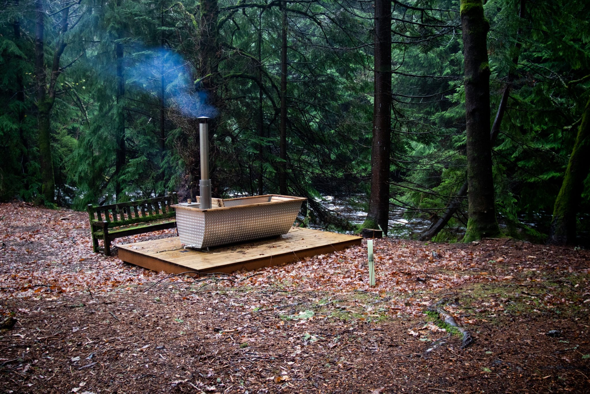 A view of the outdoor wood burning hot tub emitting smoke from its chimney shoot, with a view of the River Dye in the background between gaps in the trees.
