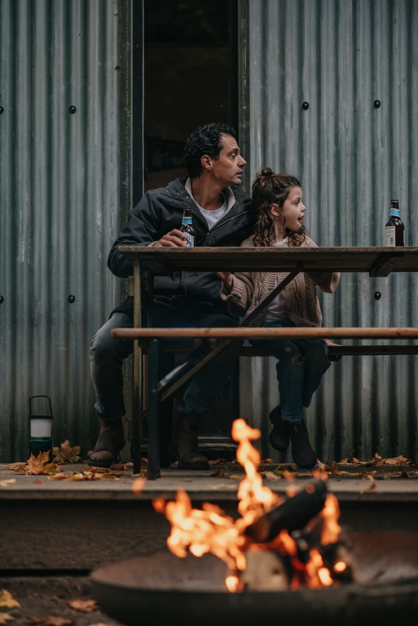 A father and daughter sat outside of a cabin with a lit fire pit in view.
