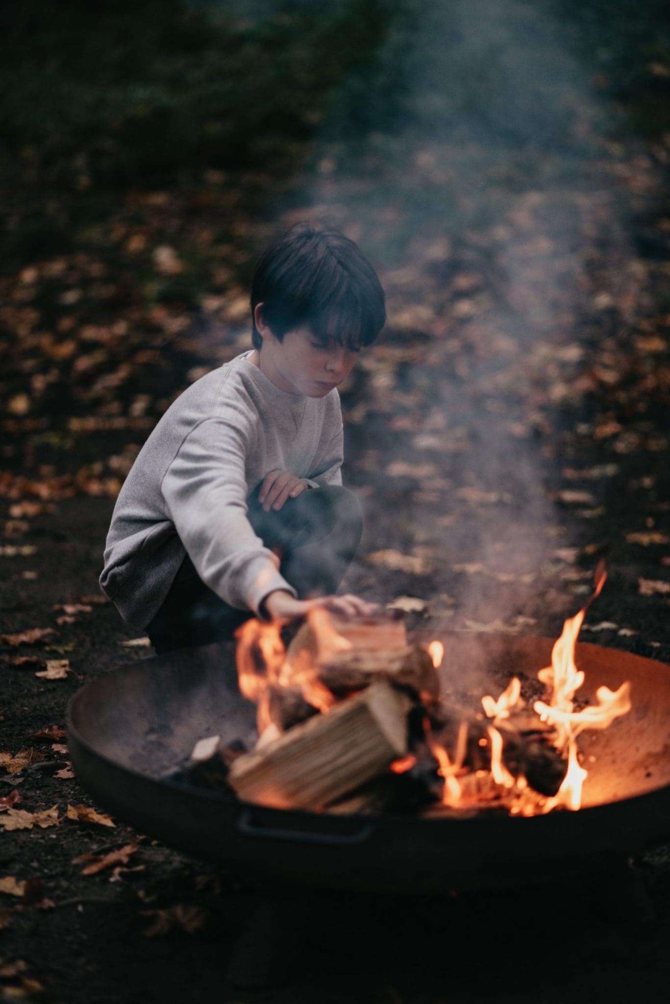 A child adding some logs to a lit fire, in an outdoor fire pit.