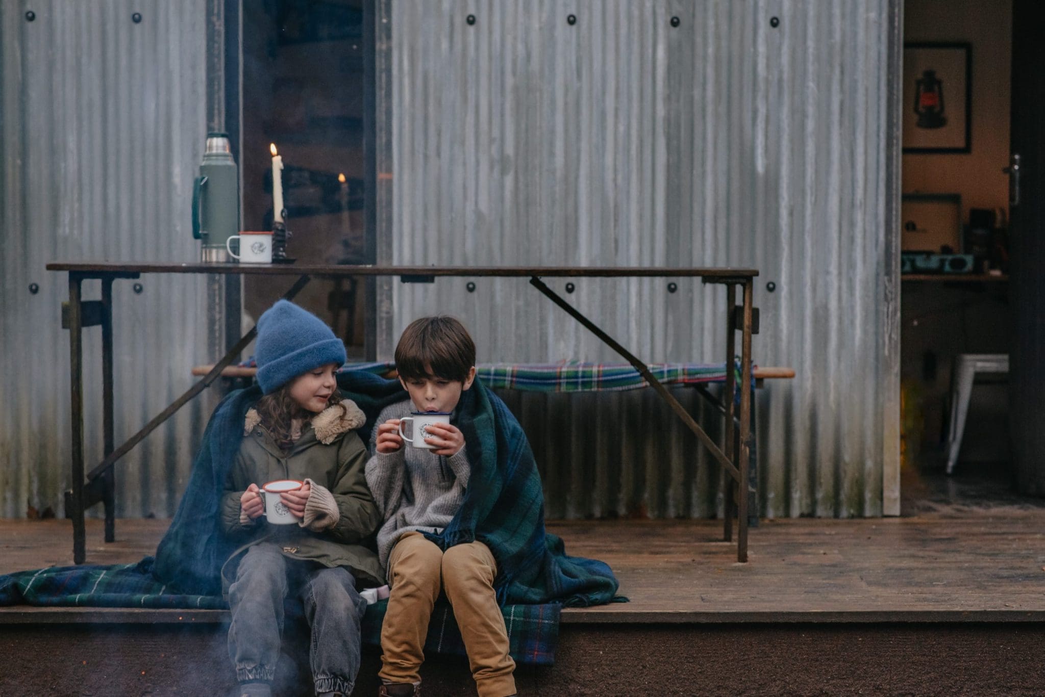 An image from the North Lodge Cabin collection, showing 2 children snuggled in a large blanket, whilst sat outside on a autumn morning.