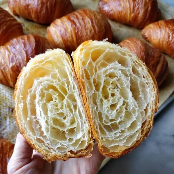 A baking tray full of freshly baked croissants, with a focus on one croissant cut in half, where the you can see its perfect layers.